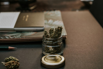 Marijuana buds in open glass jar with gold lid, notebooks and pencil on black background