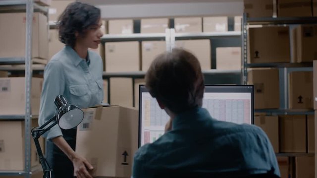 Warehouse Inventory Manager Talks to a Clerk Working with a Spreadsheet on a Personal Computer while Sitting at His Desk. In the Background Shelves Full of Cardboard Box Packages Ready For Shipping.