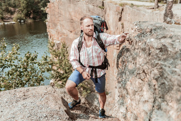 Strong hiker. Strong bearded blonde-haired hiker holding heavy backpack feeling happy while hiking near lake