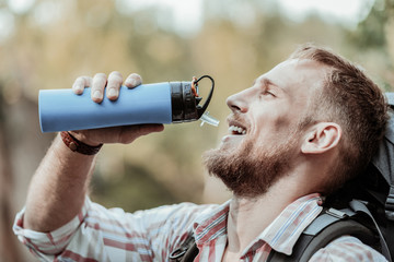 Tourist bottle. Handsome bearded dark-eyed man with heavy backpack drinking water out of his blue...