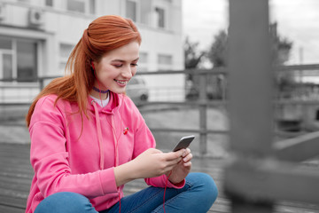 Reading jokes. Relaxed young girl sitting on the pier and smiling while looking at the screen of her modern smartphone
