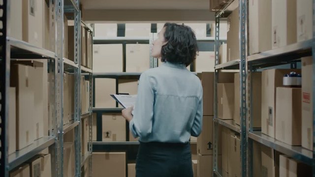 Female Inventory Manager Checks Stock, Writing in the Clipboard. Beautiful Woman Working in a Warehouse Storeroom with Rows of Shelves Full Of Cardboard Boxes, Parcels. In Slow Motion.