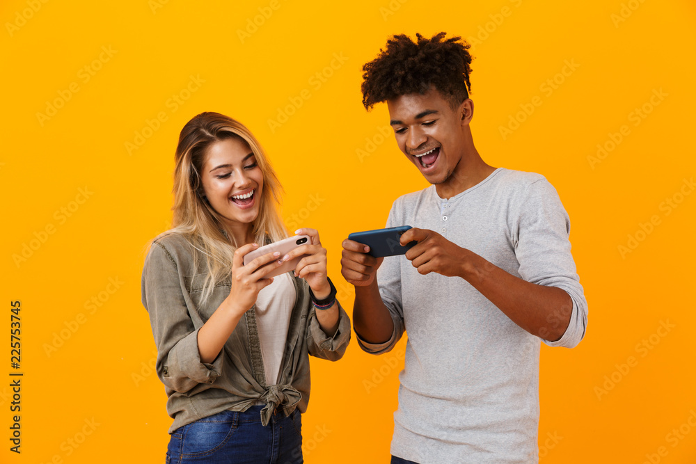 Canvas Prints Young loving couple posing isolated over yellow background play games by mobile phones.