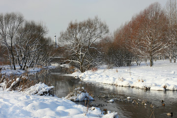 View of winter river coming among snowed rivershores and trees covered with snow and dom of church on the back