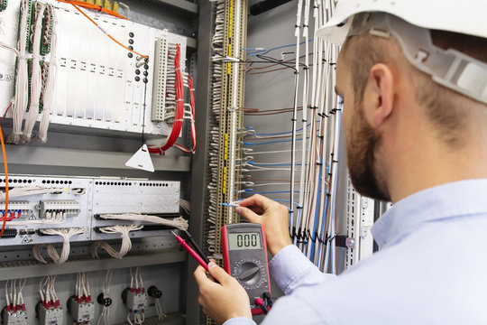 Engineer electrician with multimeter in electrical control box tests equipment. Maintenance of electrical panel. Worker in helmet in power supply cabinet. Service man is testing automation fuse box