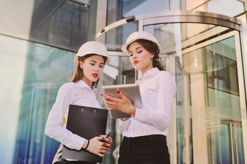 Two young cute girls in business clothes and white construction helmets discuss a business plan or contract