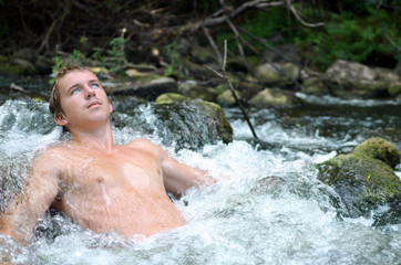 young handsome man lies in a waterfall formed by a rapids river and the water flows around his athletic body