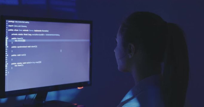 Experienced IT woman programmer works on a new program. Young woman hacker writes the source code of the software at dark room. Back view.