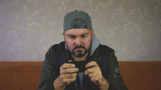City dweller. Unshaven man. Sitting on the couch and playing on the smartphone on the wall light Wallpaper. Dark clothes. Close-up portrait. Shakes his hand to the beat of music. 4K video.
