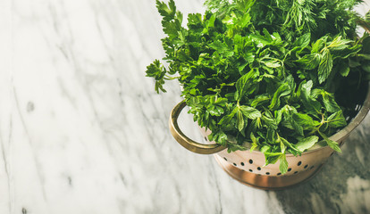 Bunch of fresh green garden herbs in brass colander over marble kitchen table background, top view,...