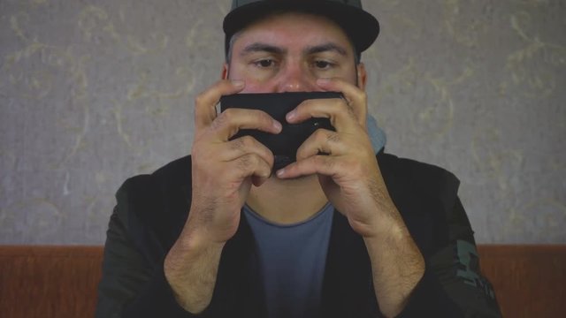 City dweller. Unshaven man. Sitting on the couch and playing on the smartphone on the wall light Wallpaper. Dark clothes. Close-up portrait. Shakes his hand to the beat of music. 4K video.