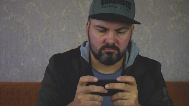 City dweller. Unshaven man. Sitting on the couch and playing on the smartphone on the wall light Wallpaper. Dark clothes. Close-up portrait. Shakes his hand to the beat of music. 4K video.