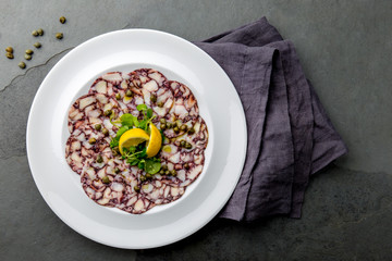 OCTOPUS CARPACCIO. Seafood Raw octopus slices with olive oil, lemon and capers on white plate. Top view. Gray stone background. © lblinova