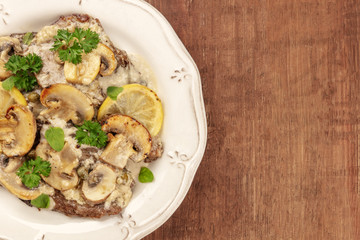 Overhead closeup photo of scallopine di vitello, veal scallopini, with a mushroom sauce, lemons, and parsley, on a dark rustic texture with copy space