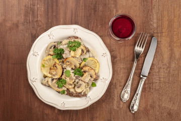 Overhead photo of scallopine di vitello, veal scallopini, with a mushroom sauce, lemons, and parsley, on a dark rustic texture with red wine and copy space