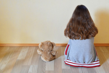 Teenage girl with teddy bear toy sitting on floor and looking at light yellow wall at empty room back view. Autism and children care and protection concept.