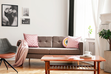 Pink pillows on sofa next to armchair in white living room interior with posters and table. Real...