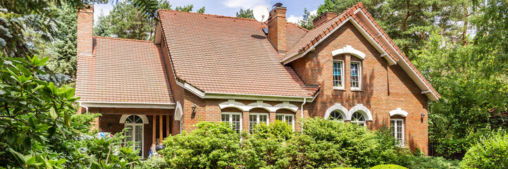 A red brick English style classic house with a steep roof and large windows surrounded by trees and...