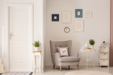 Flowers on table next to grey armchair in white living room interior with posters and door. Real...