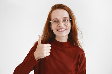 That's good idea. Portrait of emotional beautiful young red haired lady wearing maroon turtleneck and stylish glasses smiling joyfully, making thumbs up gesture as sign of approval and agreement