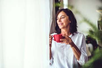 A senior woman with a cup standing by the window at home at Christmas time. - Powered by Adobe