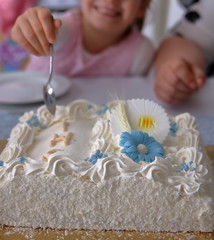 Biały tort komunijny, z niebieskimi dekoracjami, na stole, w tleręka dziewczynki, z łyżeczką,...