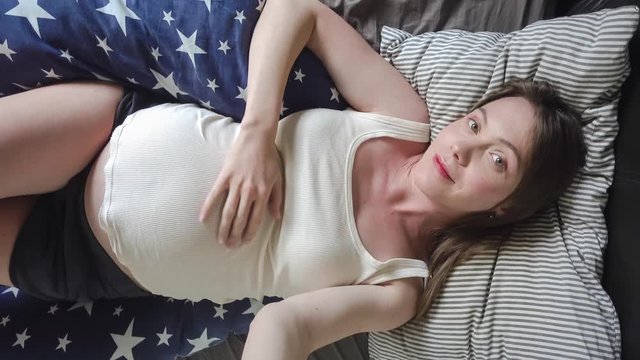 Cheerful pregnant young woman slying in bed and taking selfie with mobile phone at home top view