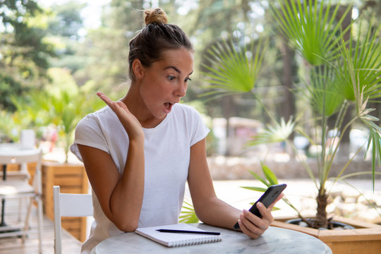 Young woman working in outdoor cafe, she got happy and surpriced by reciving salary, she looks to her smartphone with opened mouth