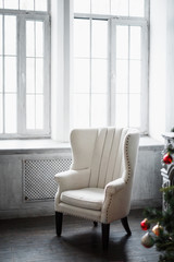 White large cozy chair in a room with gray walls and a Christmas tree in the foreground blurred. Selective focus.