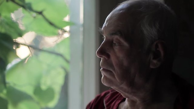 Portrait of sad lonely elderly man looking out of the window at sunset. Loneliness and depression in old age.