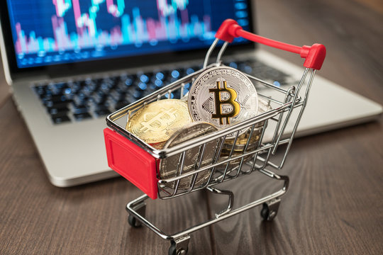 shopping trolley full of bitcoin currency with laptop showing stock market rates in the background concept on the wooden table