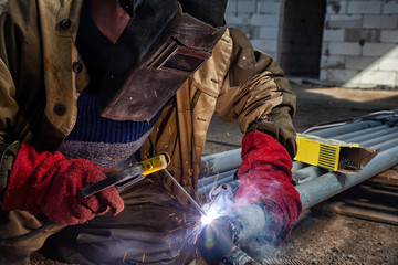 Man welder in welding mask, building uniform and red protective gloves brews metal welding machine on factory, in the background construction site - obrazy, fototapety, plakaty