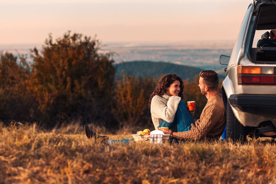 Beautiful young couple enjoying picnic time on the sunset. They drinking tea and sitting in a meadow.