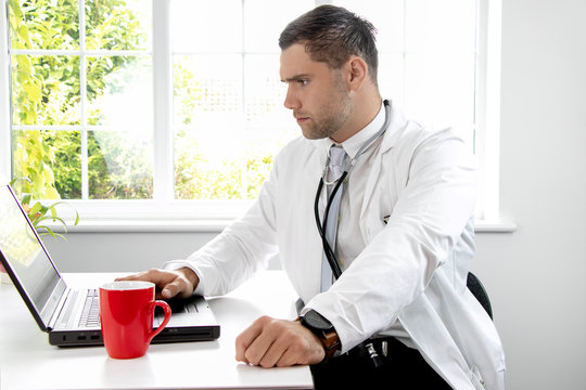 Good Looking Male Doctor In White Lab Coat Gp At Desk Looking At Laptop Computer