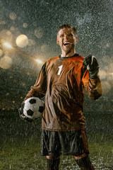 Soccer player on professional soccer night rain stadium. Dirty player in rain drops hold a football ball. Player rejoices victory © Alex