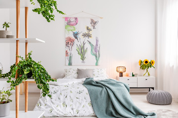 Green plants on shelves beside a bed dressed in white cotton bedding and teal blue blanket in a bright bedroom interior. Flowers and birds painted on the fabric above the bed. Real photo. - obrazy, fototapety, plakaty