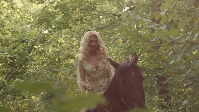 Adult pretty blonde horsewoman is riding in summer forest in sunny day. She is looking down and forward and holding reins, tilt up view