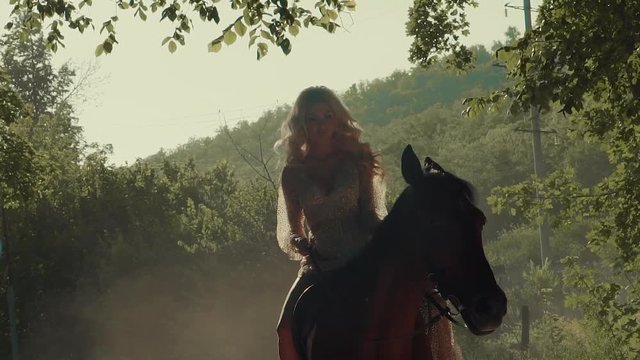 Charming young girl riding brown horse in forest in the evening wearing fantastic dress, princess. Blonde woman on ride in nature, wonderful mountain view.
