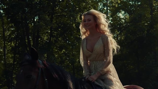 Charming blonde rider are galloping on a horse in a forest in sunny day. Her long curly hair are swaying by wind and movement, she is smiling broadly