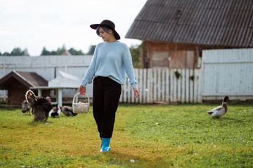 Obraz premium Young beautiful girl in a village holding a basket with chicken eggs. Autumn day, green grass, on the background of a farm. A woman is smiling in a sweater and a black hat.