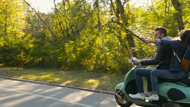 Young couple riding on their scooter at the road in the forest. Woman shooting video on the action camera, using selfiestick. View from aside. Gimbal shot.