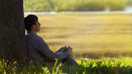 young man sitting with his back to the tree and meditating about life, the concept of religion and...