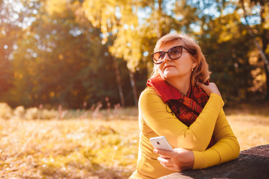 Middle-aged woman relaxing using phone outdoors. Senior lady enjoys nature in autumn forest