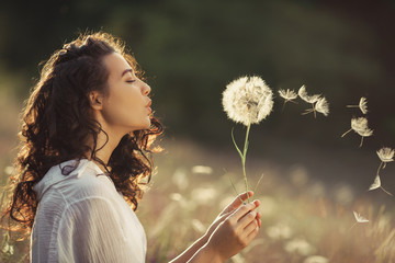 Beautiful Young Woman sitting on the field in green grass and blowing dandelion. Outdoors. Enjoy Nature. Healthy Smiling Girl on summer lawn. Allergy free concept. Gorgeous slim mixed race Caucasian - Powered by Adobe