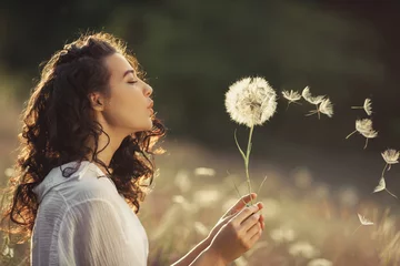 Fotobehang Beautiful Young Woman sitting on the field in green grass and blowing dandelion. Outdoors. Enjoy Nature. Healthy Smiling Girl on summer lawn. Allergy free concept. Gorgeous slim mixed race Caucasian © Buyanskyy Production