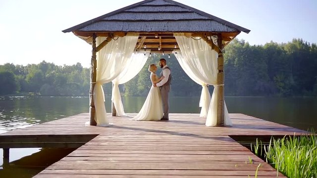 romantic pair of groom and bride in a veranda on a wooden pier of picturesque lake in summer day, approaching