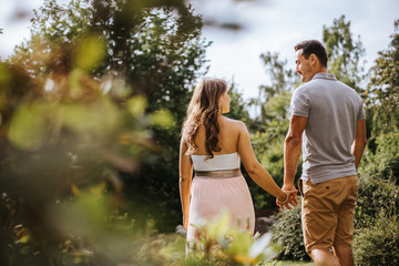Rückansicht von schöner schwangeren Frau und ihrem Partner, welche Hand in Hand durch den Park...