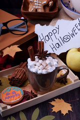 Hot chocolate with marshmallows, anise and cinnamon in a mug on a wooden tray. Halloween cookies, congratulation note and dry leaves are around