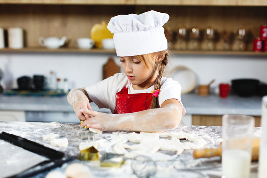 Little chief. Charming girl has fun making cookies of a dough at a cosy kitchen while her mother stands on the background