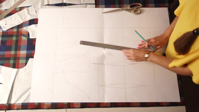 Close view of fashion designer woman hands working in studio. Caucasian female tailor drawing the layout for clothes on cloth using pencil and ruler. Atelier indoors.